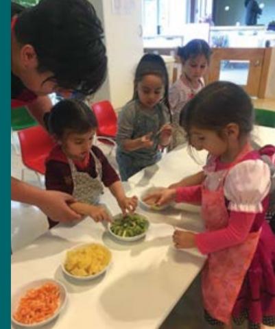 A teacher helps two students wearing aprons to take fruits from bowls on a table