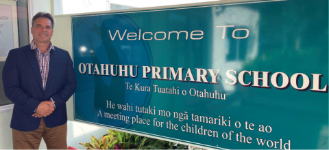 Photo of Jason Swan standing next to a sign that reads Welcome to Otahuhu Primary School, Te Kura Tuatahi o Otahuhu.