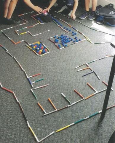 Felt pens and blocks forms a maze on a classroom floor while students' hands can be seen with a programmed Sphero ball