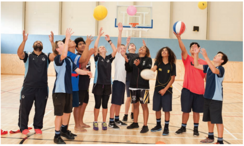 This image is a photo of a staff member and 11 students throwing balls in the air during gym time.